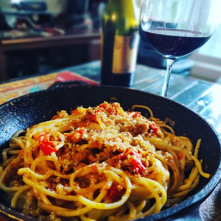 A bowl of Tomaten-Knoblauch-Pasta served with a glass of red wine and a bottle in the background.